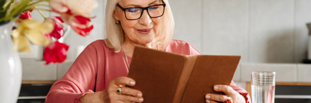 A smiling elderly woman in glasses reading a book in the light kitchen