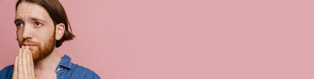 Young ginger man with beard looking aside and making pray gesture isolated over pink backgroundの写真素材