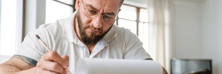 White man in eyeglasses writing down notes on clipboard while working at home