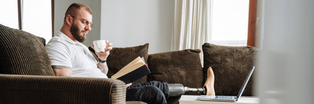 White man with prosthesis reading book and drinking coffee while siting on couch at homeの写真素材
