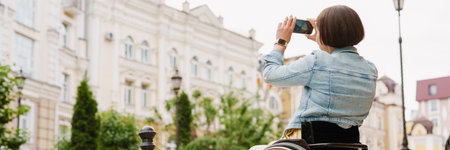 Brunette woman taking photo on mobile phone while sitting in wheelchair at city streetの写真素材