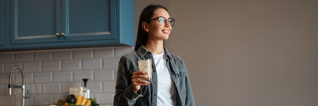 Happy mid aged brunette woman holding glass with beverage while standing at the kitchen table with...