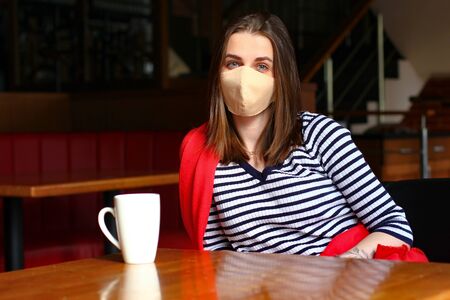 The girl in the protective medical mask is sitting in the restaurant with cup of coffeeの写真素材