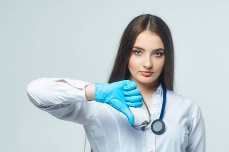 Covid19, coronavirus, a concept of healthcare and doctors. Portrait of a female doctor, shows that the bad result is a thumbs down, puts on a medical mask to prevent infection with the virus, white coatの写真素材