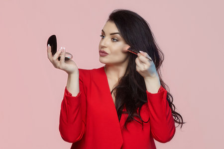charming brunette with long hair holds a compact and a brush in her hands. photo shoot on a pink...