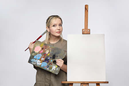 young artist poses with a palette and brush in front of an easel in the studio on a white backgroundの写真素材