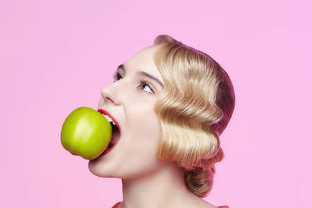 attractive young blonde with a retro hairstyle holds a green apple near her faceの写真素材