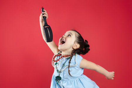 little black-haired girl plays with a comb and sings into it. funny photo shoot in the studio on a red background.の写真素材
