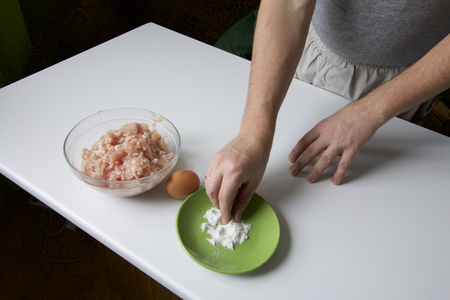 Preparing mince for meatballs. Man minced pickles and adds to it the egg. On a white table top stand: a plate with salt, egg, a container of meatの写真素材