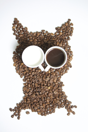 Roasted grains of black coffee. The grains are scattered in the form of an owl figurines. Owl Eyes in the form of two white cups. One of them poured brewed coffee. In another milk.の写真素材
