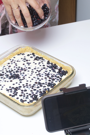 The woman adds blueberries to the pie filling. Delicate cheesecake with blueberries. On the tripod, a smartphone is installed to shoot the cooking process.の写真素材