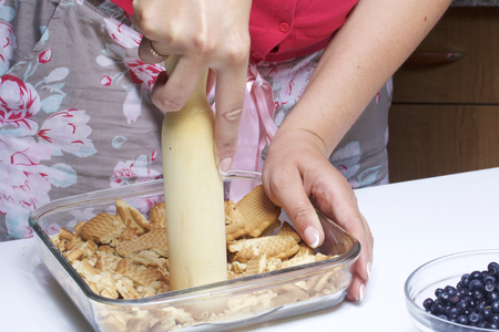 A woman is crumbling a biscuit cookie. Delicate cheesecake with blueberries. Ingredients: cookies and blueberries stand in containers on the table.の写真素材