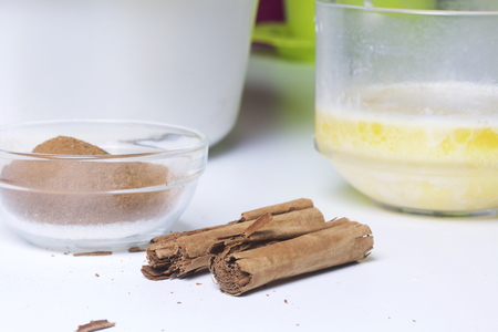 Ingredients for making pastry rolls. Crushed cinnamon and sugar are mixed in a glass container. Nearby lie cinnamon sticks and melted butter in a glass.の写真素材