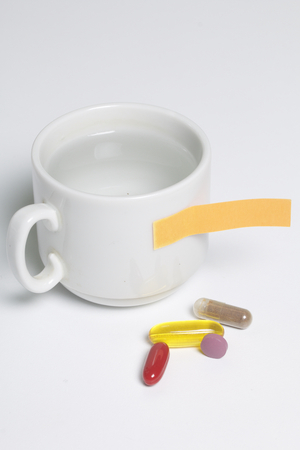 On the white surface of the table lie pills of different colors and there is a mug with water. Next is a paper sticker with a reminder.の写真素材