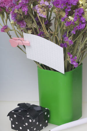 A bouquet of dried flowers is in a plastic glass. A note is attached to the flower with a clothespin. Nearby is a box with a gift. On a white background.の写真素材