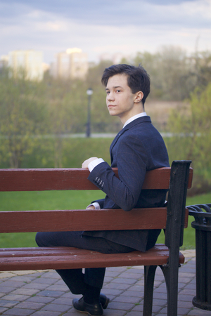A young man, a teenager, in a classic suit. Sits on a vintage bench in a spring park.の写真素材