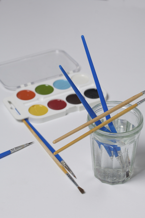 Watercolors and brushes on a white surface. Next glass for water. Subjects for creativity.の写真素材