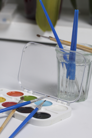Watercolors and brushes on a white surface. Next glass for water. Subjects for creativity.の写真素材