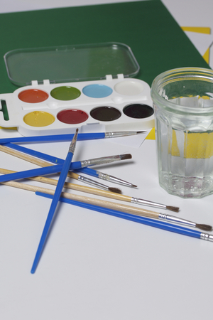 Watercolors and brushes on a white surface. Next glass for water and paper. Subjects for creativity.の写真素材