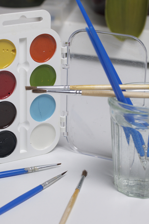 Watercolors and brushes on a white surface. Next glass for water. Subjects for creativity.の写真素材