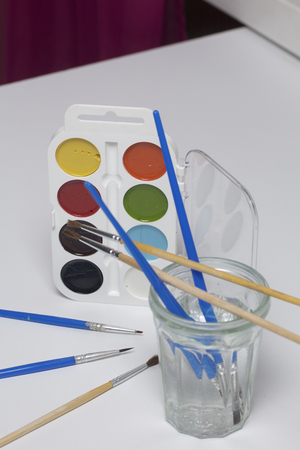 Watercolors and brushes on a white surface. Next glass for water. Subjects for creativity.の写真素材