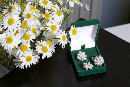 An open green velvet box for jewelry. In it lies a set: a ring and earrings with pearls. Next to the vase is a bouquet of chamomile. On a black background.の写真素材