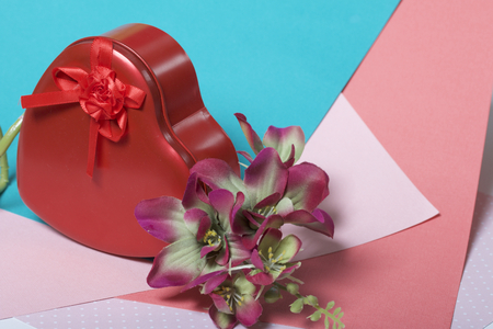 A box in the form of a red heart with a gift. Next is a flower. On a background of multicolored paper.の写真素材