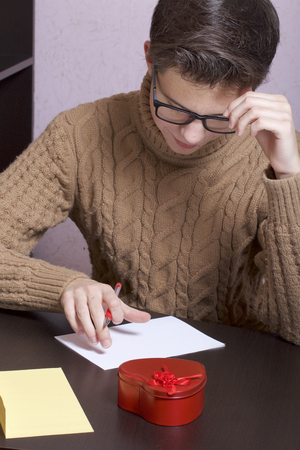 A young man writes holiday greetings on a sheet of paper. Next to the box with a gift and an envelope. Box of tin, red, made in the shape of a heart.の写真素材