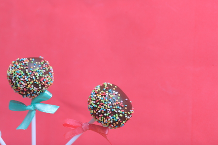 Cake Pops in chocolate with colored sprinkles. Decorated with a ribbon bow. On a red backgroundの写真素材