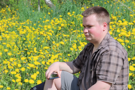A young plump man sits in a meadow with blooming dandelions. Holds a smartphone in his hand. Pensively looking in front of him.の写真素材