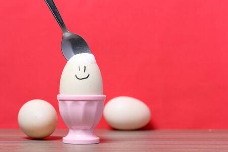 A boiled egg with a chopped shell stands in a poached. On the egg painted smiley. A spoon is stuck in it for food. Against the background of coral color.の写真素材