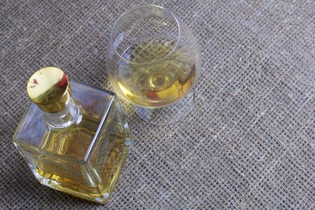 A bottle of strong alcohol. From transparent glass, a square form with a golden stopper. Near a glass with a drink poured. Against the backdrop of coarse linen fabric.の写真素材