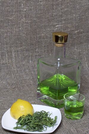 Strong tincture on tarhuna in a transparent rectangular bottle. Drink bright green poured into a glass. Next to the ingredients for cooking. Tarragon leaves and lemon.の写真素材