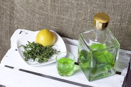 Strong tincture on tarhuna in a transparent rectangular bottle. Drink bright green poured into a glass. Next to the ingredients for cooking. Tarragon leaves and lemon.の写真素材