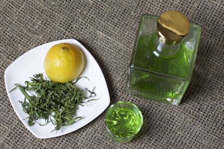 Strong tincture on tarhuna in a transparent rectangular bottle. Drink bright green poured into a glass. Next to the ingredients for cooking. Tarragon leaves and lemon.の写真素材