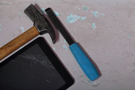 Tablet with a broken screen lies on the surface with a shabby paint. Nearby is a hammer and chisel.
