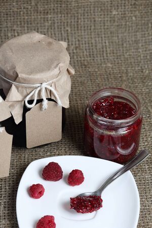 Jars of jam, covered with paper and tied with twine. On the string hang craft labels. One jar is open. Near fresh raspberries on a plate.の写真素材