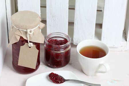 Jars of jam, covered with paper and tied with twine. On the string hang craft labels. One jar is open. Near a cup of tea.の写真素材