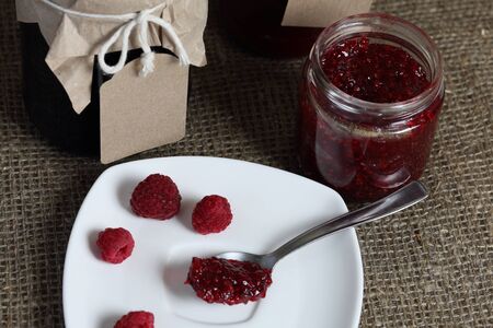 Jars of jam, covered with paper and tied with twine. On the string hang craft labels. One jar is...