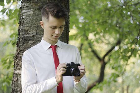 Stylishly dressed young man posing in a park. Holds the camera in his hands and works with the settings.の写真素材
