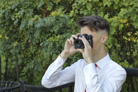 Stylishly dressed young man sits on a park bench and takes pictures. In his hands holds a SLR film...