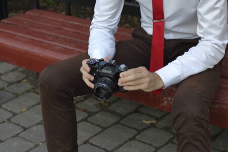 A stylishly dressed young man sits on a park bench. Stylishly dressed young man sits on a park bench. In his hands holds a SLR film camera.の写真素材
