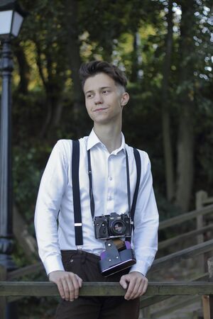 Stylishly dressed young man posing in a park. A SLR film camera hangs on the neck.