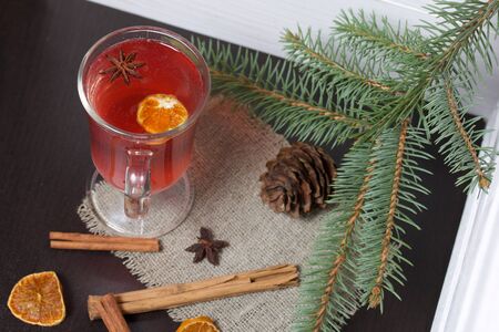 A glass of red drink stands on a piece of linen. A slice of dried orange and anise floats in it. Nearby are cinnamon sticks and dried oranges and a spruce branch.の写真素材