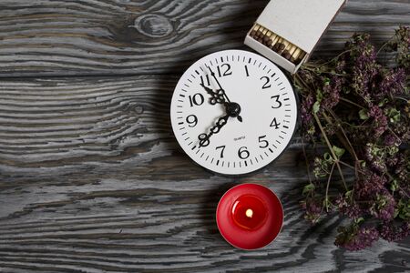 Burning candle on brushed boards. Near the clock and a bouquet of thyme.
