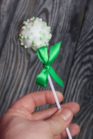 A man holds cake pops in white glazed chocolate with pink sprinkles. A green bow is tied on a stick. Against the background of brushed pine boards painted in black and white.の写真素材