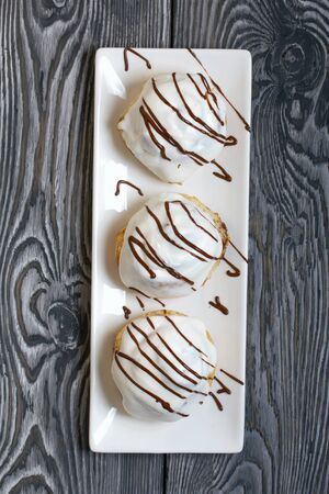 Cinnabons in white glaze, decorated with chocolate. Lie on a plate. Against the background of...