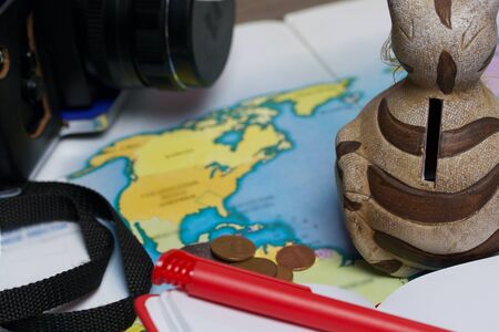 A ceramic piggy bank with a slot for coins is on the world map. Several coins are scattered nearby, a camera, a notebook and a pen lie. Piggy bank for travel.の写真素材