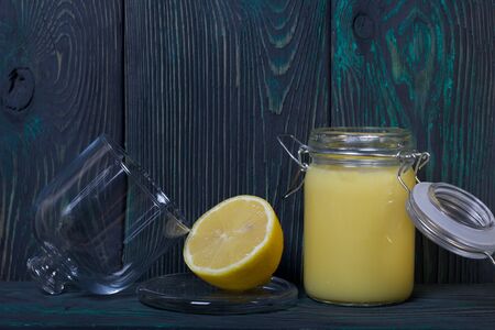 Lemon Kurd in a glass jar. Nearby is half a lemon in a container with a glass lid. Against the background of brushed boards.の写真素材