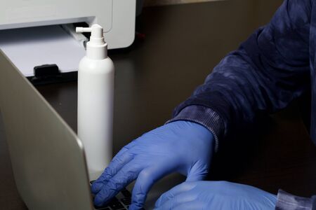 A man in protective rubber gloves works at a laptop. Nearby is a disinfectant. Work in self-isolation during a global epidemic.の写真素材